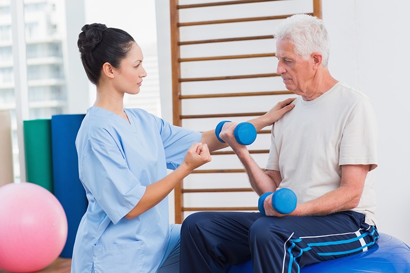 Physical Therapist working with elderly man during outpatient visit on movement therapy with small hand weights.