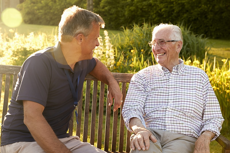 Man Visiting Senior Male Relative on a sunny outdoor bench In Assisted Living Facility